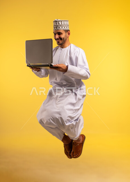إنجاز المهام المطلوبة عبر اللابتوب، استخدام جهاز تقني حديث ومتطور، بورتريه لرجل عربي خليجي عماني يرتدي الدشداشة والكمة يقفز عاليا يستخدم الحاسوب المحمول، مفهوم ادارة الأعمال عن بعد، دمج التكنولوجيا بالعمل، الجسم بالطول الكامل، خلفية صفراء