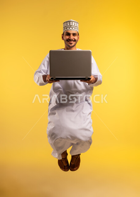 دمج التكنولوجيا بالعمل، إنجاز المهام المطلوبة عبر اللابتوب، استخدام جهاز تقني حديث ومتطور، بورتريه لرجل عربي خليجي عماني يرتدي الدشداشة والكمة يقفز عاليا يستخدم الحاسوب المحمول، مفهوم ادارة الأعمال عن بعد، الجسم بالطول الكامل، خلفية صفراء