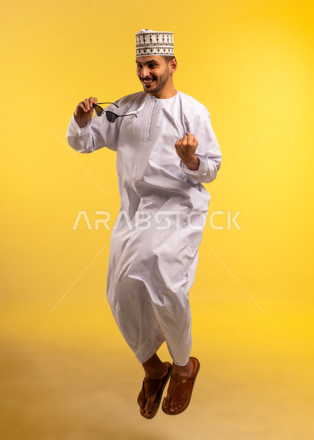 Caring about appearance, the concept of elegance and handsomeness, portrait of a young Arab Gulf Omani man wearing a dishdasha and a turban, holding sunglasses in his hand, jumping high with gestures of self-confidence and pleasure, full-length body, yellow background