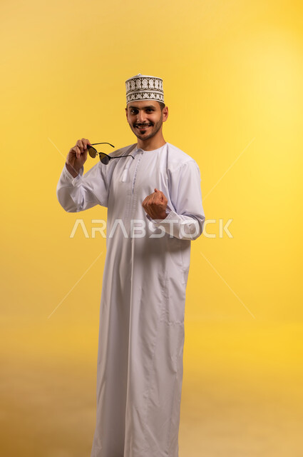 The concept of elegance and handsomeness, a portrait of a young Arab Gulf Omani man wearing a dishdasha and a kummah, holding sunglasses in his hand, looking at the camera with gestures of self-confidence and pleasure, interest in outward appearance, yellow background