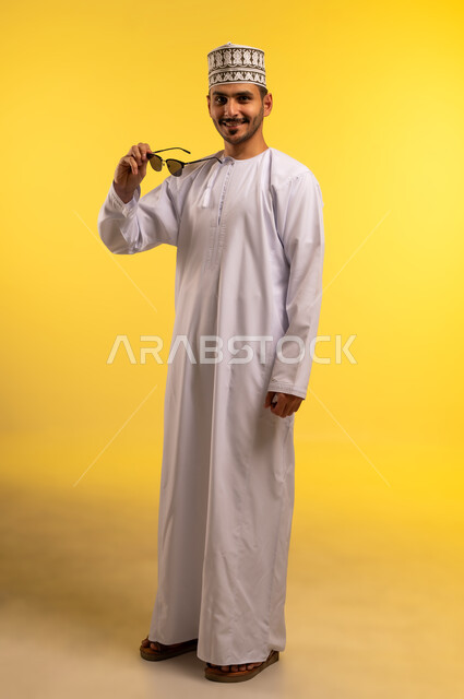الاهتمام بالمظهر الخارجي، مفهوم الأناقة والوسامة، بورتريه لشاب عربي خليجي عماني يرتدي الدشداشة والكمة يمسك بيده النظارة الشمسية ينظر إلى الكاميرا بايماءات الثقة بالنفس والسرور, الجسم بالطول الكامل، خلفية صفراء