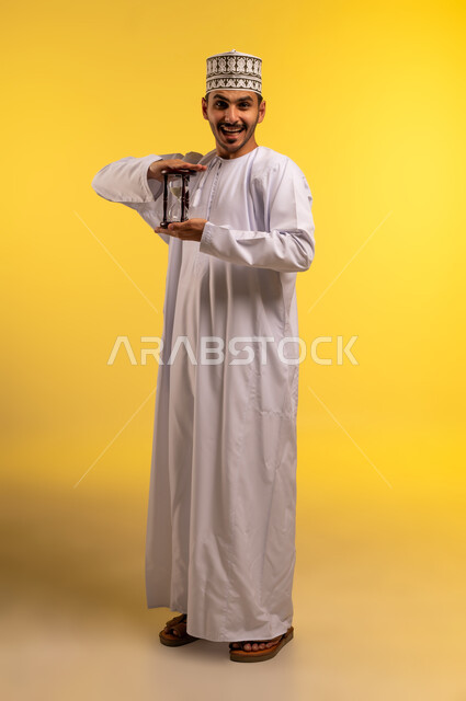 مفهوم إدارة الأعمال وتنظيم الجدول الزمني, بورتريه لشاب عربي خليجي عماني يرتدي الدشداشة والكمة يمسك بيده الساعة الرملية الزجاجية (البنكام) ينظر إلى الكاميرا بايماءات الفرح والسرور, اهمية مراقبة الوقت, إدراك قيمة اللحظات الثمينة, الجسم بالطول الكامل، خلفية صفراء