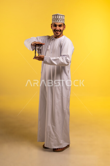 اهمية مراقبة الوقت, مفهوم إدارة الأعمال وتنظيم الجدول الزمني, بورتريه لشاب عربي خليجي عماني يرتدي الدشداشة والكمة يمسك بيده الساعة الرملية الزجاجية (البنكام) ينظر إلى الكاميرا بايماءات الفرح والسرور, إدراك قيمة اللحظات الثمينة, الجسم بالطول الكامل، خلفية صفراء
