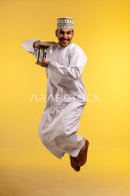 إدراك قيمة اللحظات الثمينة, مفهوم إدارة الأعمال وتنظيم الجدول الزمني, بورتريه لشاب عربي خليجي عماني يرتدي الدشداشة والكمة يمسك بيده الساعة الرملية الزجاجية (البنكام) يقفز عاليا ينظر إلى الكاميرا بايماءات السرور, اهمية مراقبة الوقت, الجسم بالطول الكامل، خلفية صفراء