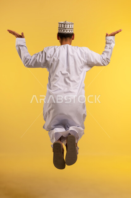 القفز عاليا بتعابير الفرح والحماس، بورتريه من الخلف لشاب عربي خليجي عماني يرتدي الدشداشة والكمة يرفع يديه بإيماءات السرور والسعادة, الاهتمام بالمظهر الخارجي، مفهوم الرجولة والأناقة، الجسم بالطول الكامل، خلفية صفراء