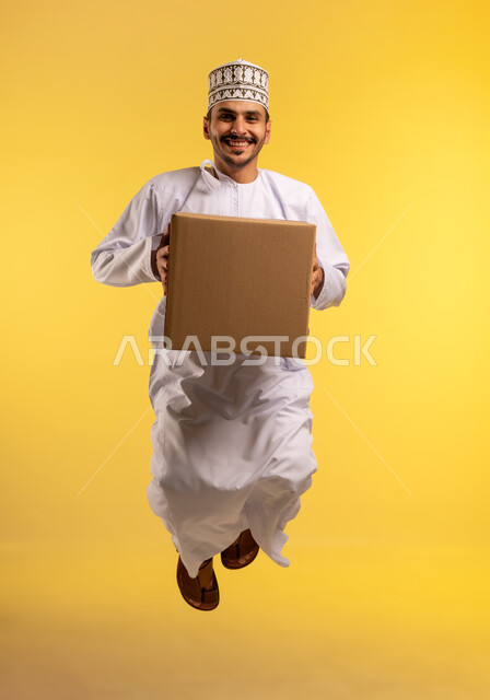 خدمة الشحن والتوصيل ذات الجودة العالية، التسوق وطلب المنتجات عبر الإنترنت، توصيل الطلبات في الوقت المحدد وبشكل سليم، بورتريه لشاب عربي خليجي عماني يرتدي الدشداشة والكمة يقفز عاليا يحمل صندوق كرتوني، شراء الاحتياجات المنزلية، الجسم بالطول الكامل، خلفية صفراء