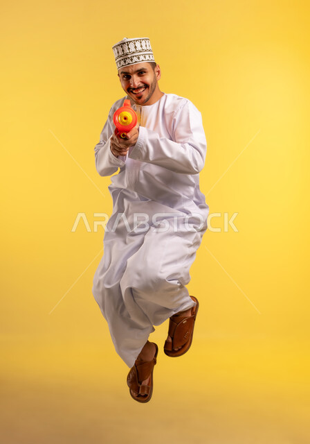 قضاء وقت ممتع بالألعاب المائية في فصل الصيف، بورتريه لشاب عربي خليجي عماني يرتدي الدشداشة والكمة يحمل مسدس مائي بلاستيكي يقوم باللعب بإيماءات السرور والسعادة، القفز عاليا بتعابير الحماس والفرح، مفهوم الترفيه عن النفس والتسلية، الجسم بالطول الكامل، خلفية بيضاء