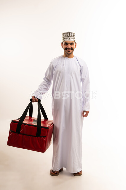 أداء العمل بدقة واتقان، التسوق وشراء الحاجيات عبر الانترنت، ضمان سلامة الطرود المرسلة، بورتريه لشاب عربي خليجي عماني يرتدي الدشداشة والكمة يحمل حقيبة حمراء ينظر إلى الكاميرا بإيماءات السرور، عملية الشحن والتوصيل الموثوقة، الجسم بالطول الكامل، خلفية بيضاء