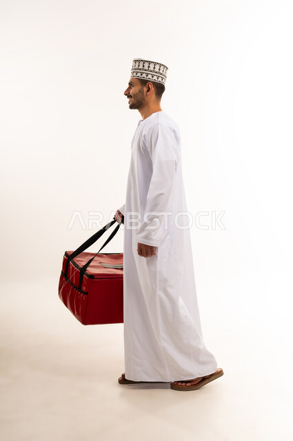 Walking and going somewhere, shopping and buying necessities online, ensuring the safety of the parcels sent, side portrait of a young Arab Gulf Omani man wearing a dishdasha and a kummah carrying a red bag, reliable shipping and delivery process, performing work with precision and mastery, full body, white background