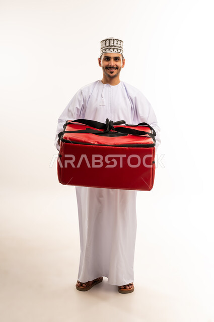 عملية الشحن والتوصيل الموثوقة، أداء العمل بدقة واتقان، التسوق وشراء الحاجيات عبر الانترنت، ضمان سلامة الطرود المرسلة، بورتريه لشاب عربي خليجي عماني يرتدي الدشداشة والكمة يحمل حقيبة حمراء ينظر إلى الكاميرا بإيماءات السرور، الجسم بالطول الكامل، خلفية بيضاء