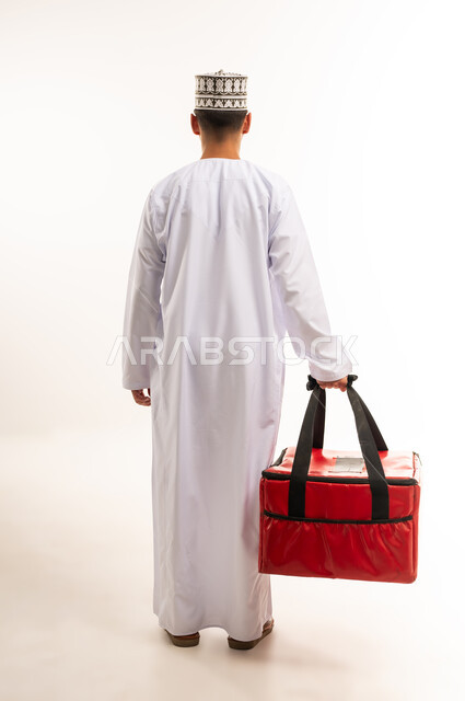 التسوق وشراء الحاجيات عبر الانترنت، ضمان سلامة الطرود المرسلة، بورتريه من الخلف لشاب عربي خليجي عماني يرتدي الدشداشة والكمة يحمل حقيبة حمراء، عملية الشحن والتوصيل الموثوقة، أداء العمل بدقة واتقان، الجسم بالطول الكامل، خلفية بيضاء