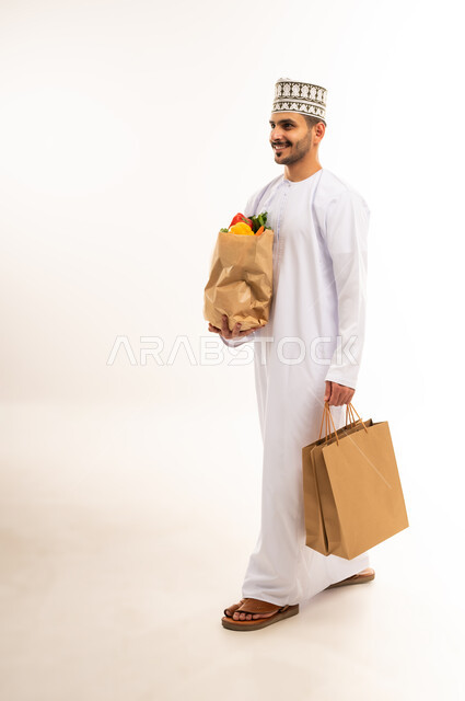 المشي والذهاب إلى مكان ما، التبضع واقتناء الخضراوات والفاكهة، توفير واحضار المواد الغذائية، بورتريه لشاب عربي خليجي عماني يرتدي الدشداشة والكمة يحمل مجموعة أكياس وكيس ورقي مليء بالخضار والفواكه، التسوق وشراء الحاجيات المنزلية، الجسم بالطول الكامل، خلفية بيضاء