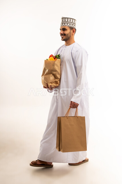 توفير واحضار المواد الغذائية، المشي والذهاب إلى مكان ما، التبضع واقتناء الخضراوات والفاكهة، بورتريه لشاب عربي خليجي عماني يرتدي الدشداشة والكمة يحمل مجموعة أكياس وكيس ورقي مليء بالخضار والفواكه، التسوق وشراء الحاجيات المنزلية، الجسم بالطول الكامل، خلفية بيضاء