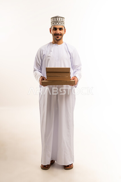 التسوق وطلب المنتجات عبر الإنترنت، توصيل الطلبات في الوقت المحدد وبشكل سليم، خدمة الشحن و التوصيل ذات الجودة العالية، بورتريه لشاب عربي خليجي عماني يرتدي الدشداشة والكمة يحمل بيده صندوق كرتوني مغلق، شراء الاحتياجات المنزلية، الجسم بالطول الكامل، خلفية بيضاء