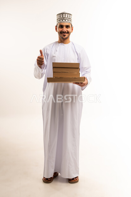 علامة الجودة والامتياز، توصيل الطلبات في الوقت المحدد، خدمة الشحن والتوصيل ذات الجودة العالية، بورتريه لشاب عربي خليجي عماني يرتدي الدشداشة والكمة يحمل صندوق كرتوني يرفع اصبع الابهام بإيماءات الاعجاب، التسوق وطلب المنتجات عبر الإنترنت، الجسم بالطول الكامل، خلفية بيضاء