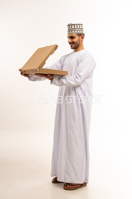 توصيل الطلبات في الوقت المحدد وبشكل سليم، خدمة الشحن و التوصيل ذات الجودة العالية، بورتريه لشاب عربي خليجي عماني يرتدي الدشداشة والكمة يحمل بيده صندوق كرتوني، التسوق وطلب المنتجات عبر الإنترنت، شراء الاحتياجات المنزلية، الجسم بالطول الكامل، خلفية بيضاء