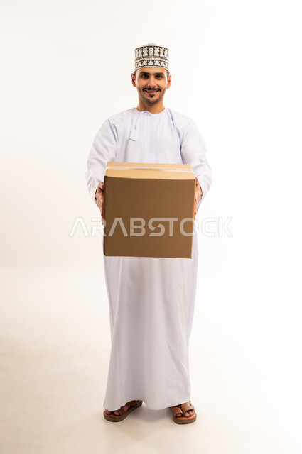 High quality shipping and delivery service, online shopping and ordering, on time and accurate delivery, portrait of a young Omani Gulf Arab man wearing a dishdasha and a turban holding a closed cardboard box, buying household necessities, full body, white background