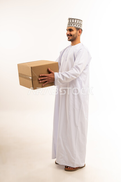 التسوق وطلب المنتجات عبر الإنترنت، توصيل الطلبات في الوقت المحدد وبشكل سليم، خدمة الشحن و التوصيل ذات الجودة العالية، بورتريه لشاب عربي خليجي عماني يرتدي الدشداشة والكمة يحمل بيده صندوق كرتوني مغلق، شراء الاحتياجات المنزلية، الجسم بالطول الكامل، خلفية بيضاء