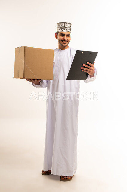خدمة الشحن و التوصيل ذات الجودة العالية، التسوق وطلب المنتجات عبر الإنترنت، توصيل الطلبات في الوقت المحدد وبشكل سليم، بورتريه لشاب عربي خليجي عماني يرتدي الدشداشة والكمة يحمل بيده صندوق كرتوني مغلق، شراء الاحتياجات المنزلية، الجسم بالطول الكامل، خلفية بيضاء