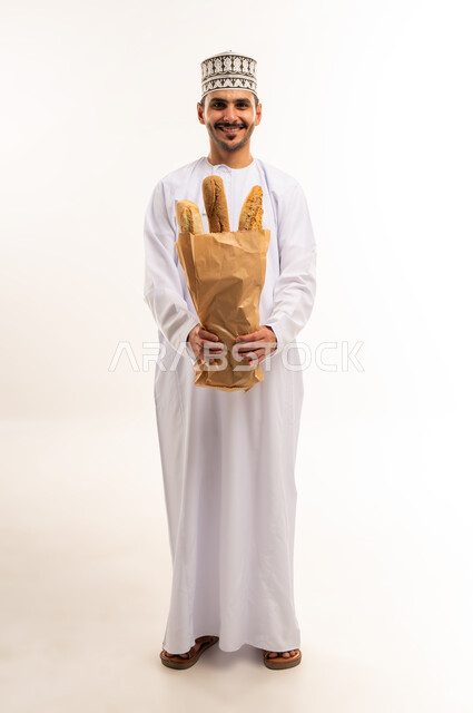 شراء الاحتياجات المنزلية والمواد الغذائية المختلفة، المعجنات الطازجة الشهية، بورتريه لشاب عربي خليجي عماني يرتدي الدشداشة والكمة يحمل كيس من الخبز ينظر إلى الكاميرا بإيماءات السرور، اهمية العناصر الغذائية الموجودة في المخبوزات، الجسم بالطول الكامل، خلفية بيضاء