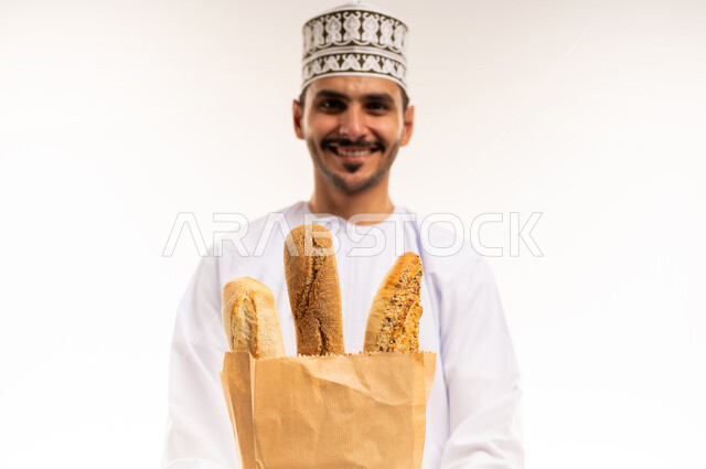 اهمية العناصر الغذائية الموجودة في المخبوزات، المعجنات الطازجة الشهية، بورتريه مقرب لشاب عربي خليجي عماني يرتدي الدشداشة والكمة يحمل كيس من الخبز ينظر إلى الكاميرا بإيماءات السرور، شراء الاحتياجات المنزلية والمواد الغذائية المختلفة، خلفية بيضاء