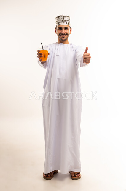 علامة الجودة والامتياز، فاكهة منعشة ولذيذة، اتباع نظام غذائي صحي، بورتريه لشاب عربي خليجي عماني يرتدي الدشداشة والكمة يمسك كوب من عصير البرتقال الطازج يرفع اصبع الابهام بإيماءات القبول والاعجاب، المحافظة على سلامة الجهاز المناعي، الجسم بالطول الكامل، خلفية بيضاء