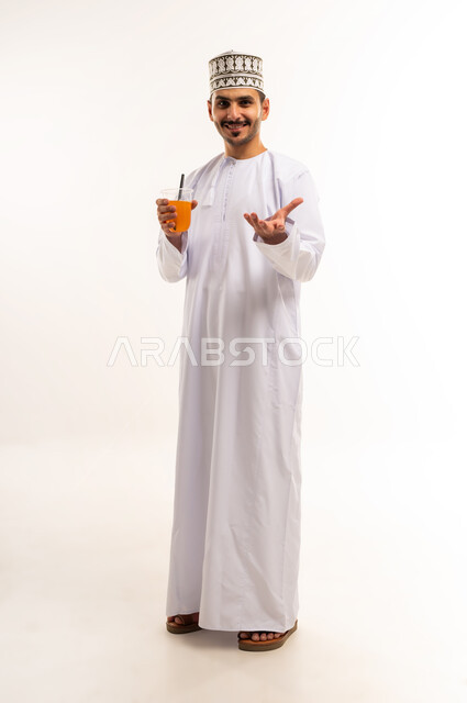المحافظة على سلامة الجهاز المناعي، مفهوم العناية والاهتمام بالصحة, فاكهة منعشة ولذيذة، اتباع نظام غذائي صحي، بورتريه لشاب عربي خليجي عماني يرتدي الدشداشة والكمة يمسك كوب من عصير البرتقال الطازج ينظر إلى الكاميرا بإيماءات السرور، الجسم بالطول الكامل، خلفية بيضاء