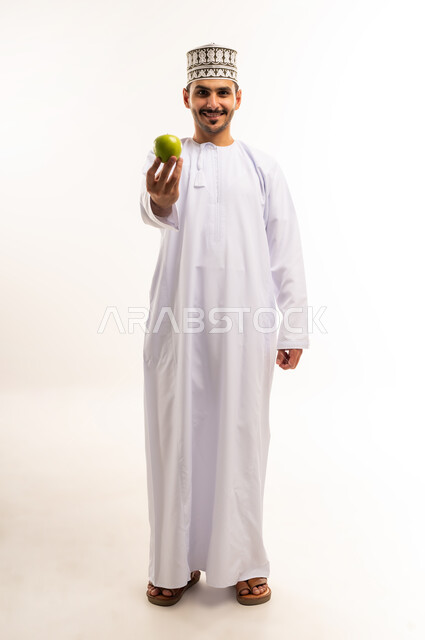 اتباع نظام غذائي، فاكهة طبيعية وطازجة, أهمية العناصر الغذائية الموجودة في التفاح, الطعام الصحي والمتوازن, بورتريه لشاب عربي خليجي عماني يرتدي الدشداشة والكمة يمسك تفاحة ينظر إلى الكاميرا بإيماءات السرور والسعادة، وجبة خفيفة لذيذة، الجسم بالطول الكامل، خلفية بيضاء