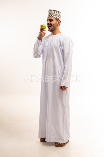 فاكهة طبيعية وطازجة, أهمية العناصر الغذائية الموجودة في التفاح, الطعام الصحي والمتوازن, بورتريه لشاب عربي خليجي عماني يرتدي الدشداشة والكمة يمسك تفاحة يقوم بأكلها، اتباع نظام غذائي، وجبة خفيفة لذيذة، الجسم بالطول الكامل، خلفية بيضاء