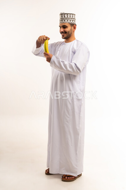 Importance of nutrients in bananas, healthy and balanced food, portrait of a young Arab Gulf Omani man wearing a dishdasha and a kimono holding a banana getting ready to eat it, diet, fresh and natural fruit, delicious snack, full body, white background