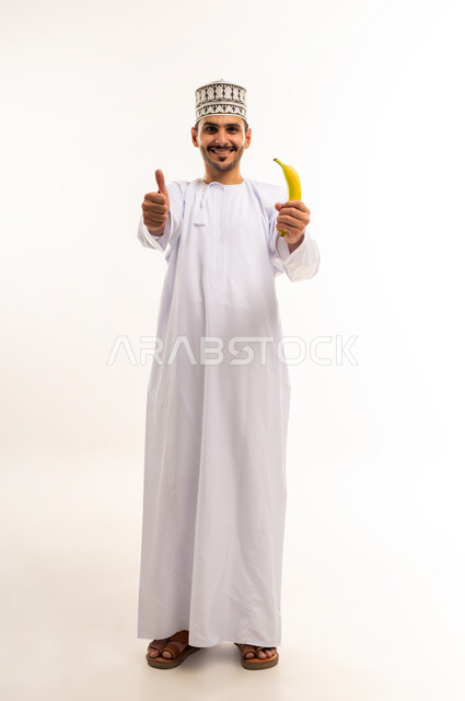 علامة الجودة والامتياز، أهمية العناصر الغذائية الموجودة في الموز, الطعام الصحي والمتوازن, بورتريه لشاب عربي خليجي عماني يرتدي الدشداشة والكمة يمسك موزة يرفع اصبع الابهام بإيماءات القبول والاعجاب، اتباع نظام غذائي، فاكهة طبيعية وطازجة, الجسم بالطول الكامل، خلفية بيضاء