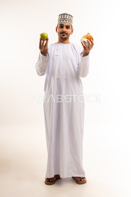 المقارنة بين الطعام الصحي و الغير صحي، تناول الوجبات السريعة, بورتريه لرجل عربي خليجي عماني مبتسم يرتدي الدشداشة والكمه يحمل بيده شطيرة البرغر والتفاحة ينظر الى الكاميرا بإيماءات الحيرة والتردد، صورة الجسم بالطول الكامل، خلفية بيضاء