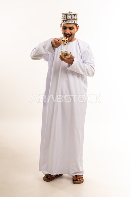 تعابير الفرح والسرور والاعجاب, بورتريه لرجل عربي خليجي عماني مبتسم يرتدي الدشداشة والكمه يحمل بيده سلطة الفواكه يتناول الطعام الصحي الشهي، صورة الجسم بالطول الكامل، خلفية بيضاء