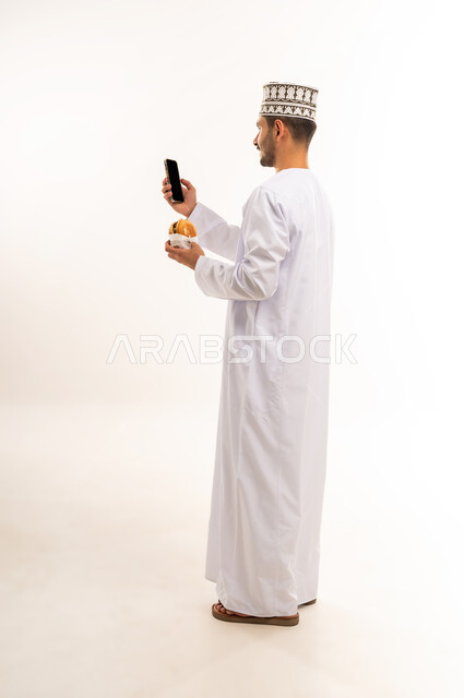 طلب الأطعمة عبر تطبيقات الجوال, طلب واستلام الدليفري, بورتريه من الجانب لرجل عربي خليجي عماني مبتسم يرتدي الدشداشة والكمه يحمل بيده الهاتف المحمول و شطيرة البرغر، عرض شاشة سوداء فارغة، صورة الجسم بالطول الكامل، خلفية بيضاء