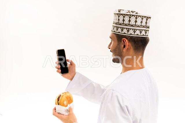 التقاط صورة السيلفي التذكارية، طلب الأطعمة عبر تطبيقات الجوال, طلب واستلام الدليفري, بورتريه مقرب من الجانب لرجل عربي خليجي عماني مبتسم يرتدي الدشداشة والكمه يحمل بيده الهاتف المحمول و شطيرة البرغر، عرض شاشة سوداء فارغة، خلفية بيضاء