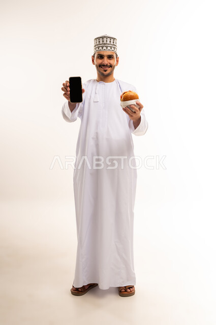 طلب واستلام الدليفري, طلب الأطعمة عبر تطبيقات الجوال, بورتريه لرجل عربي خليجي عماني مبتسم يرتدي الدشداشة والكمه يحمل بيده الهاتف المحمول و شطيرة البرغر ينظر الى الكاميرا بإيماءات السرور، عرض شاشة سوداء فارغة، صورة الجسم بالطول الكامل، خلفية بيضاء