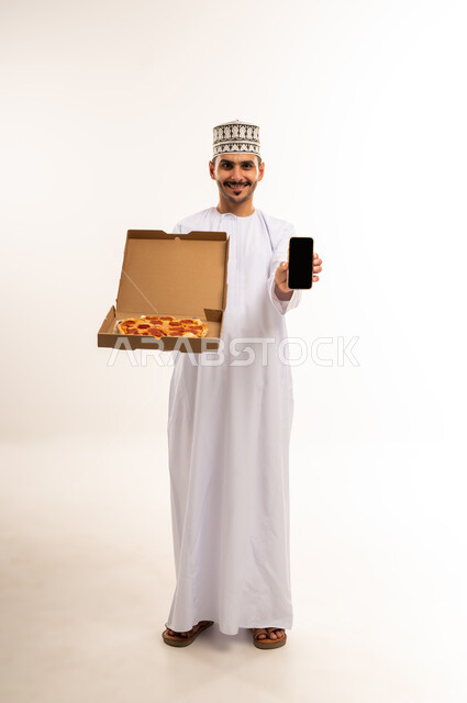 Ordering food via mobile applications, portrait of a smiling Omani Gulf Arab man wearing a dishdasha and a kummah holding a mobile phone and a delicious pizza pie looking at the camera with gestures of pleasure, ordering and receiving delivery, displaying a blank black screen, full-length body image, white background