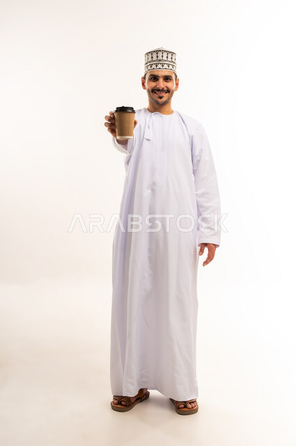تأثير الكافيين على نشاط الدماغ والذاكرة، النظر الى الكاميرا بإيماءات السرور، الاستمتاع بتناول المشروبات الساخنة والأعشاب الشتوية الدافئة، بورتريه لرجل عربي خليجي عماني يرتدي الدشداشة والكمه يحمل بيده كوب ورقي مقوى من القهوة، صورة الجسم بالكامل، خلفية بيضاء