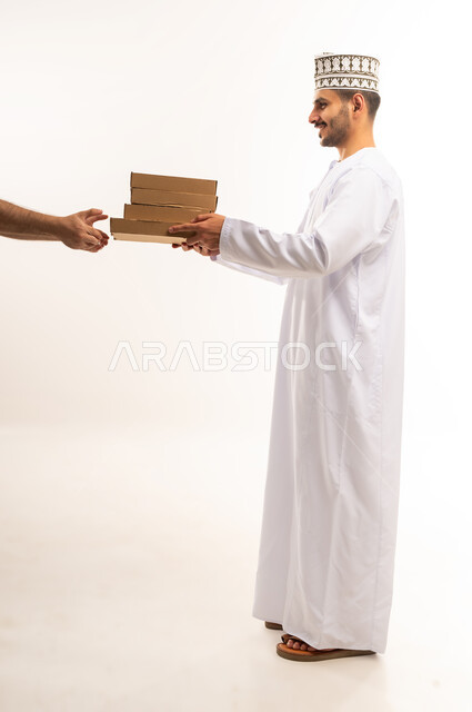 خدمات الشحن ذات الجودة العالية, طلب المنتجات عبر الانترنت، عملية التوصيل الآمنة والموثوقة, بورتريه لشاب عربي خليجي عماني يرتدي الكمه و الدشداشة يقوم بتسليم الطلبات للزبائن، توصيل الطلبيات بالوقت المحدد و بشكل سليم, صورة الجسم بالطول الكامل, خلفية بيضاء