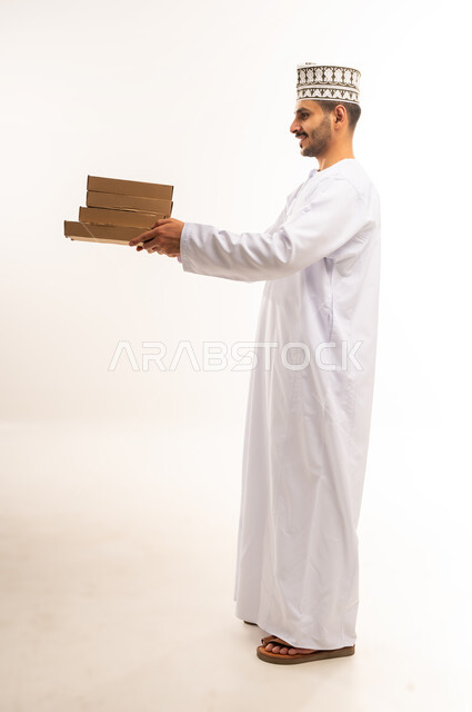 طلب المنتجات عبر الانترنت، عملية التوصيل الآمنة والموثوقة, بورتريه لشاب عربي خليجي عماني يرتدي الكمه و الدشداشة يقوم بتسليم الطلبات للزبائن، خدمات الشحن ذات الجودة العالية, توصيل الطلبيات بالوقت المحدد و بشكل سليم, صورة الجسم بالطول الكامل, خلفية بيضاء