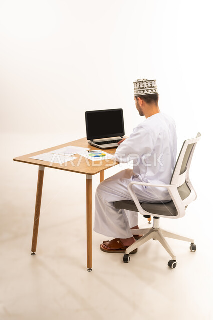 بيانات خسائر وأرباح الشركة, تطوير استراتيجية العمل وخطة النمو، مراقبة الاقتصاد والمعلومات المالية، بورتريه من الخلف لرجل عربي خليجي عماني يرتدي الكمه و الدشداشة يجلس على كرسي أمام طاولة خشبية يعمل على جهاز الحاسوب المحمول, خلفية بيضاء