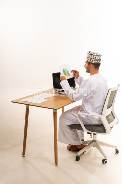 Business strategy development and growth plan, monitoring economy and financial information, financial and economic investment, side portrait of an Arab Gulf Omani man wearing a kimono and a dishdasha sitting on a chair in front of a wooden table studying and analyzing data, company profit and loss data, white background