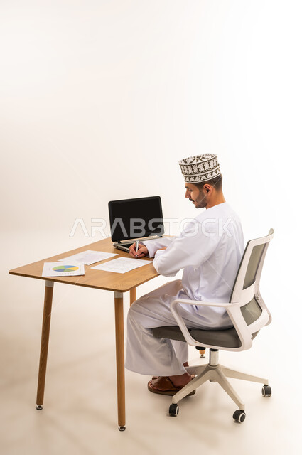 التحقق والتدقيق في المعلومات, دمج التقنية الحديثة المتطورة في الحياة العملية,بورتريه لرجل عربي خليجي عماني يرتدي الكمه و الدشداشة يجلس على كرسي أمام طاولة خشبية يقوم بتوقيع التقرير الشهري, مهنة ووظيفه شبابية عمانية, خلفية بيضاء