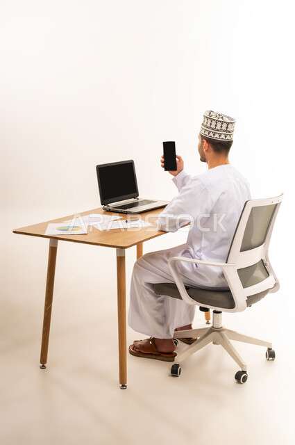 دمج التقنية الحديثة المتطورة مع الحياة العملية, التسويق الرقمي و ترويجات إعلانية, عرض شاشة سوداء فارغه عبر الحاسوب المحمول, بورتريه مقرب من الخلف لرجل عربي خليجي عماني يرتدي الزي التقليدي يحمل بيده جهاز الجوال, خلفية بيضاء