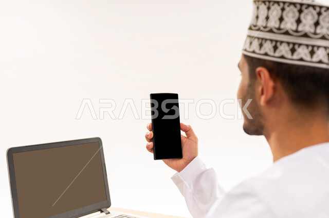 التسويق الرقمي و ترويجات إعلانية, عرض شاشة سوداء فارغه عبر الحاسوب المحمول, بورتريه مقرب من الخلف لرجل عربي خليجي عماني يرتدي الزي التقليدي يحمل بيده جهاز الجوال, دمج التقنية الحديثة المتطورة مع الحياة العملية, خلفية بيضاء