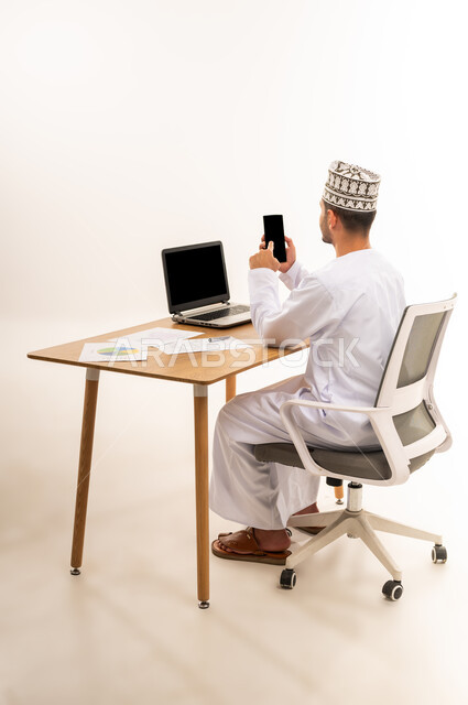 دمج التقنية الحديثة المتطورة مع الحياة العملية, عرض شاشة سوداء فارغه عبر الحاسوب المحمول, بورتريه من الخلف لرجل عربي خليجي عماني يرتدي الزي التقليدي يجلس على كرسي أمام طاولة خشبية يضغط باصبع السبابة على شاشة الجوال, خلفية بيضاء