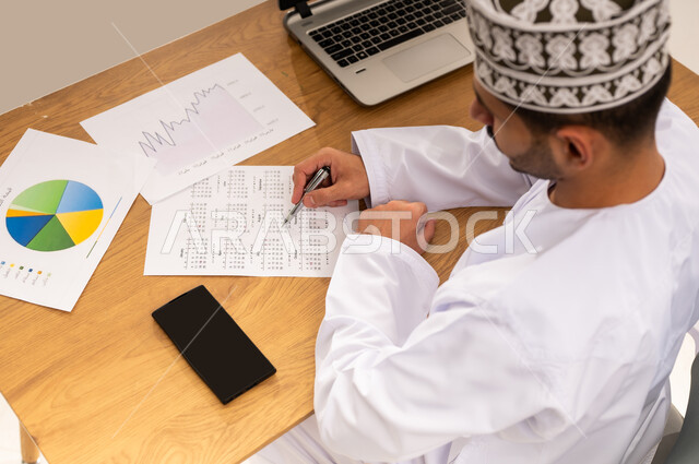 قسم التحليلات والإحصائيات البيانية، تدوين الملاحظات و النتائج, متابعة مخطط الرسم البياني للنمو الاقتصادي، بورتريه  مقرب من الاعلى لموظف عربي خليجي عماني يجلس على كرسي أمام طاولة خشبية يرتدي الكمه و الدشداشة يقوم بإنجاز المهام اليومية ضمن مقر العمل، خلفية بيضاء