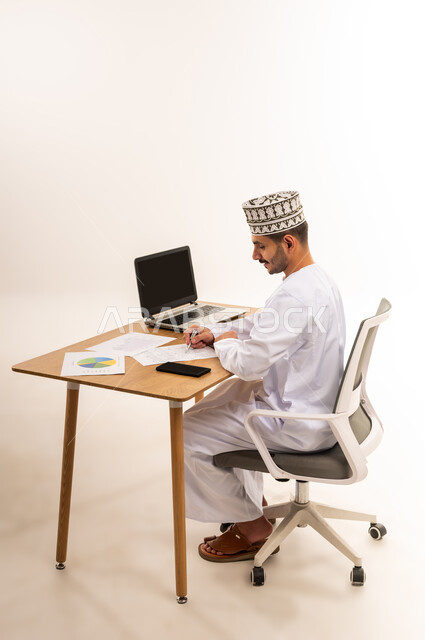 Recording and recording notes and results, following up on the economic growth chart, portrait of an Arab Gulf Omani employee sitting on a chair in front of a wooden table wearing a kemeh and a dishdasha, completing daily tasks within the workplace, the analysis and graphic statistics department, white background