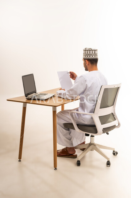 التدقيق على التقرير الشهري, القيام بأداء المهام اليومية المكتبيه, بورتريه من الخلف لرجل عربي خليجي عماني يرتدي الكمه و الدشداشة يجلس على الكرسي أمام طاولة خشبية يحمل بيده قلم وورقة يشير الى شيء محدد, مهنة ووظيفة شبابية, خلفية بيضاء
