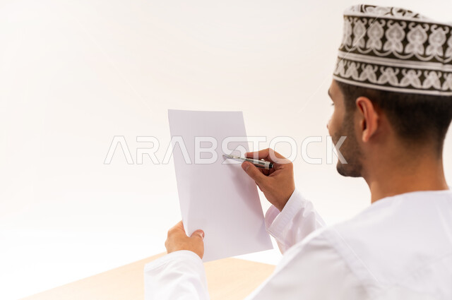 القيام بأداء المهام اليومية المكتبيه, بورتريه مقرب من الخلف لرجل عربي خليجي عماني يرتدي الكمه و الدشداشة يحمل بيده قلم وورقة يشير الى شيء محدد, مهنة ووظيفة شبابية, التدقيق على التقرير الشهري, خلفية بيضاء